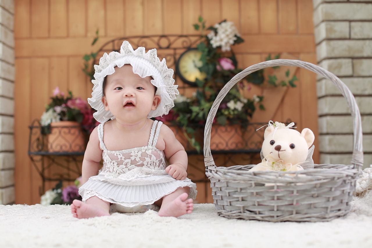 a baby sitting next to a basket with a teddy bear, a picture, by David Diao, pexels, white lace clothing, high definition photo, young goddess, panels