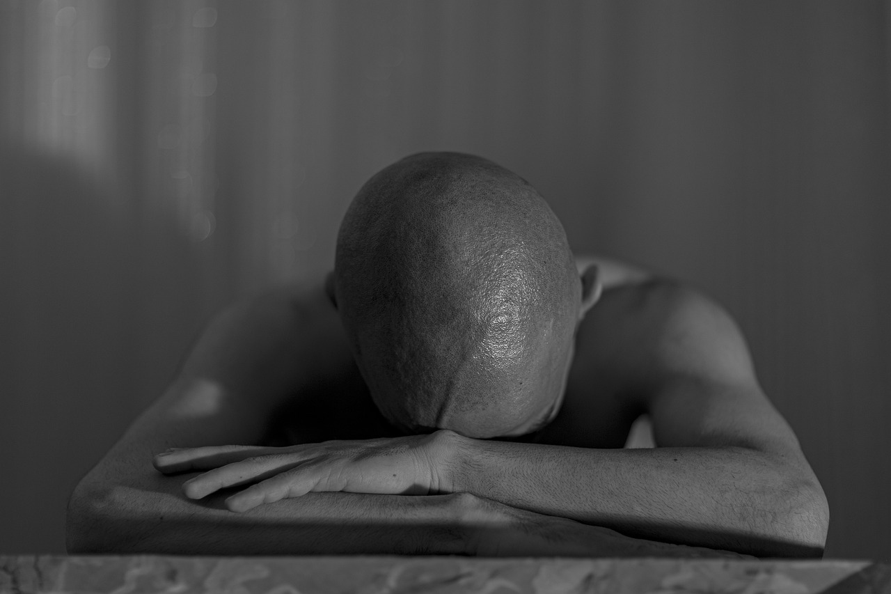 a man sitting at a table with his head on his hands, a black and white photo, inspired by Robert Mapplethorpe, unsplash, hyperrealism, shaved bald head, padmasana, resting after a hard fight, portrait of saitama