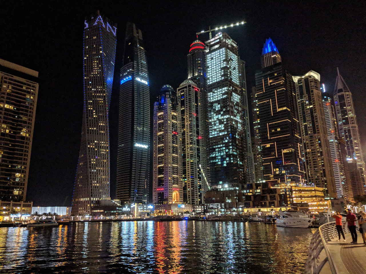 a group of people standing on a dock next to a body of water, hurufiyya, night life buildings, with tall glass skyscrapers, view from the sea, luxury lifestyle