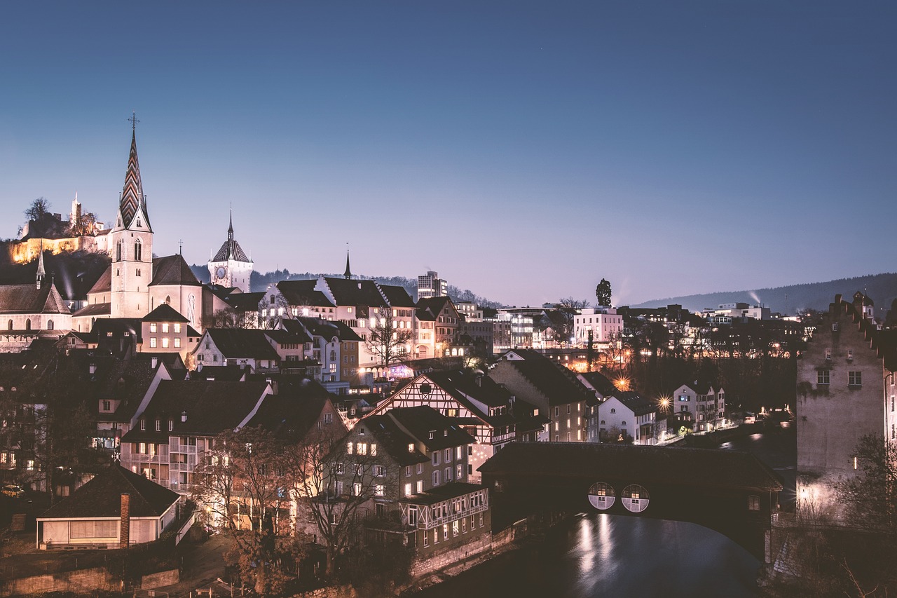 a view of a city lit up at night, by Jakob Gauermann, shutterstock, grossmünster, faded glow, twilight ; wide shot, white buildings with red roofs