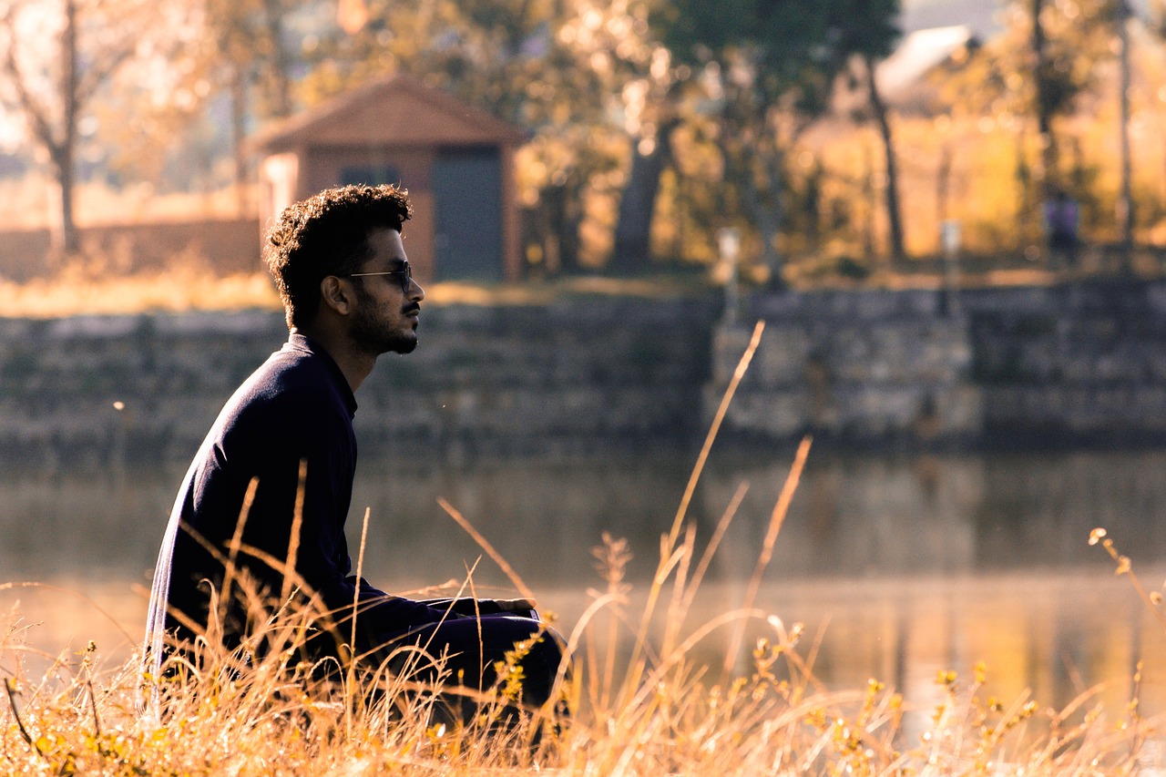 a man sitting on top of a grass covered field, a picture, pexels, romanticism, brown skinned, gazing at the water, relaxed. gold background, sitting at a pond
