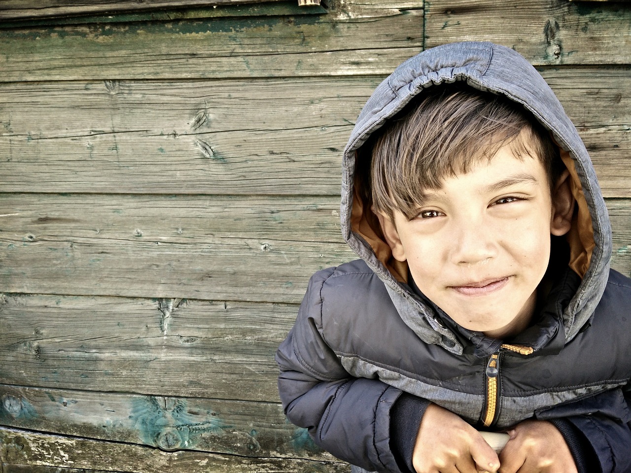 a young boy standing in front of a wooden wall, a picture, pexels, background is a slum, smiling spartan, cold grey eyes, childish