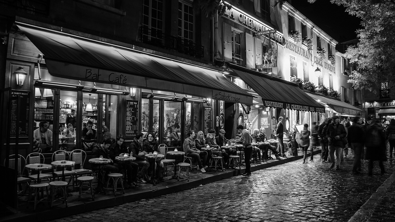 a group of people sitting outside of a restaurant at night, a black and white photo, by Raphaël Collin, pexels, fine art, cobblestone streets, in the early morning, the city of paris, by rainer hosch