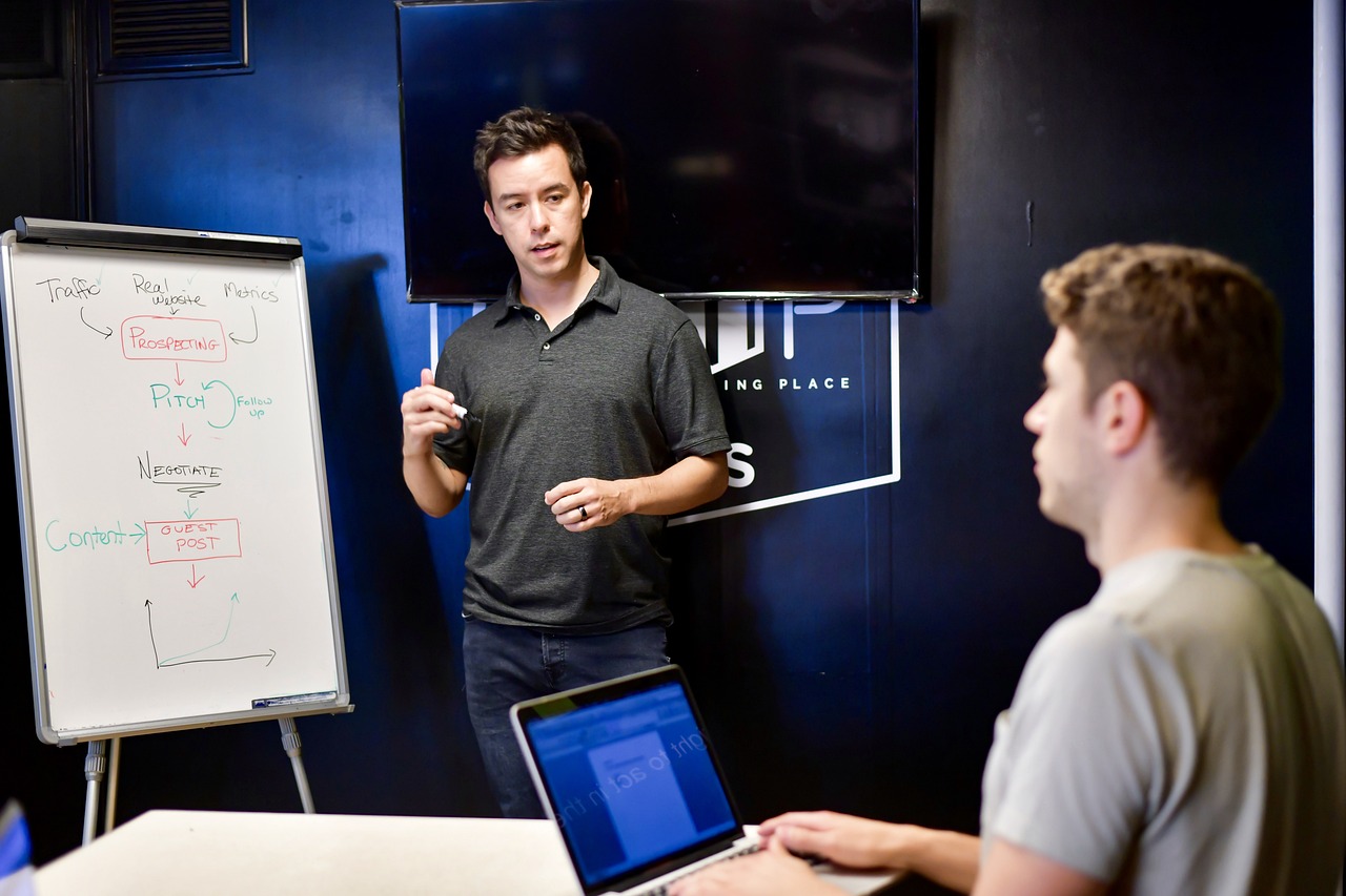 a man giving a presentation to a group of people, a photo, josh black, working on a laptop at a desk, blackboard in background, high res photo