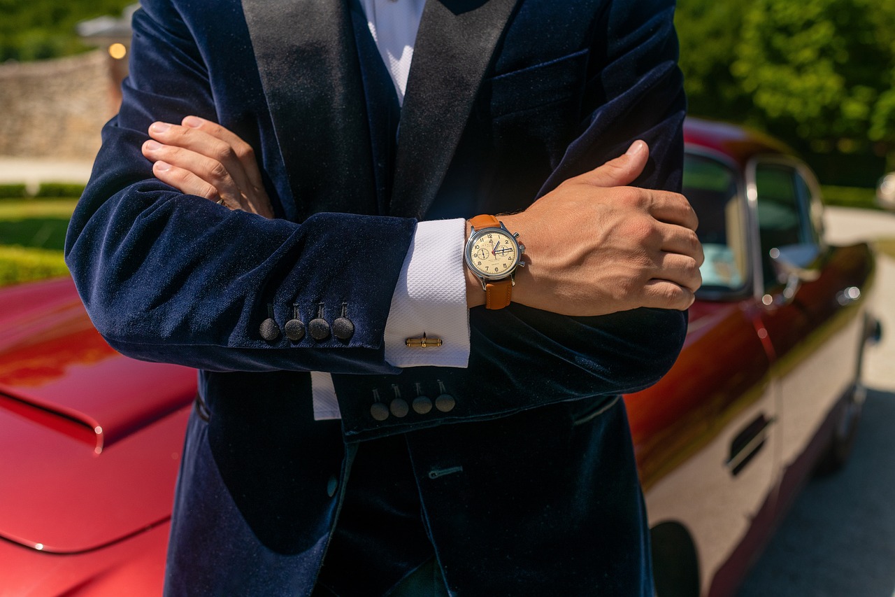 a man in a suit standing in front of a red car, inspired by Harry Haenigsen, unsplash, renaissance, gold watch, ocher details, leather cuffs around wrists, blue gold suit