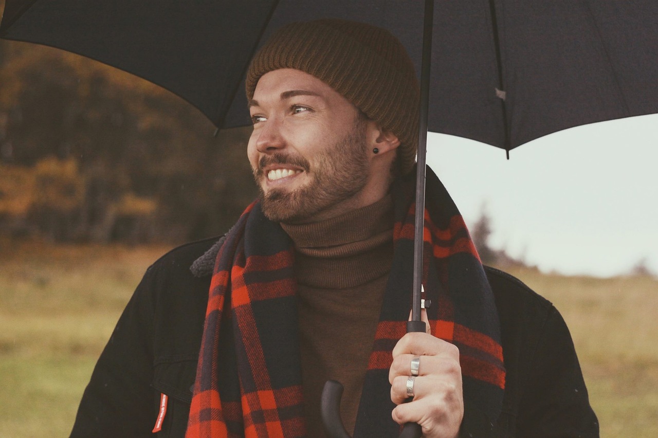 a man holding an umbrella in a field, a photo, inspired by Hamish MacDonald, unsplash, wearing turtleneck, stubble beard, tartan scarf, he is wearing a hat