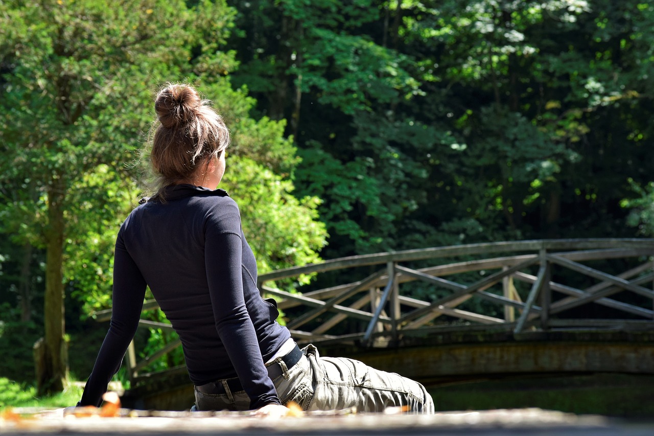 a woman sitting on the ground in front of a bridge, a picture, by Dietmar Damerau, sunny day in the forrest, with his back turned, zen feeling, with a park in the background