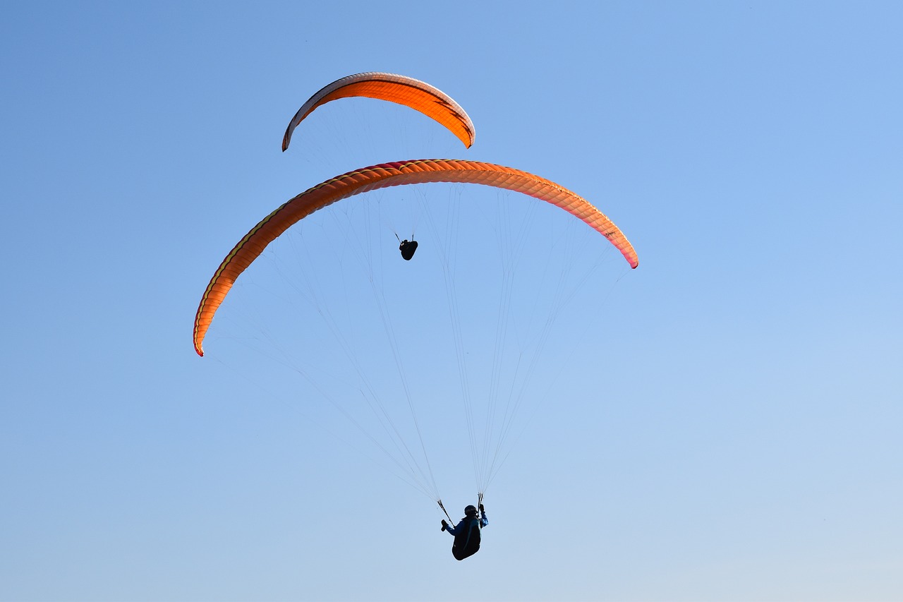 a couple of people that are parasailing in the sky, a picture, figuration libre, 7 0 mm photo, strong blue and orange colors, hard morning light, narrow wings behind