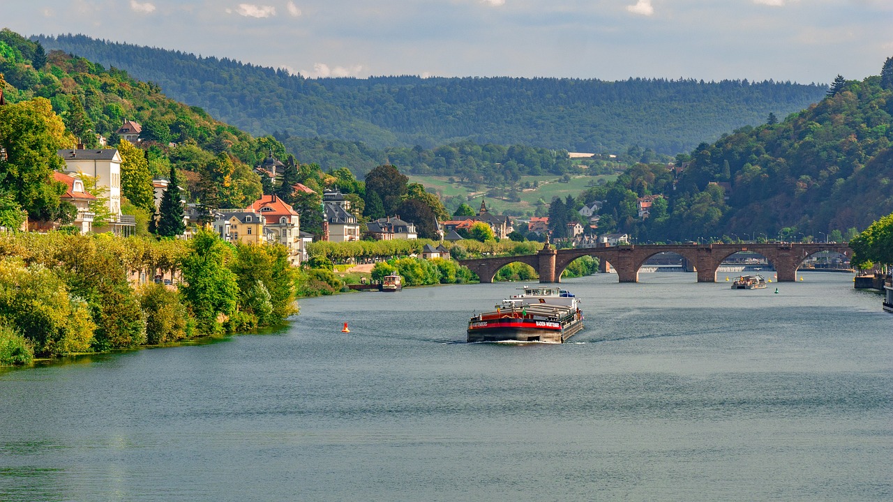 a boat traveling down a river next to a bridge, a photo, by Werner Gutzeit, shutterstock, hills in the background, germany. wide shot, red river, port