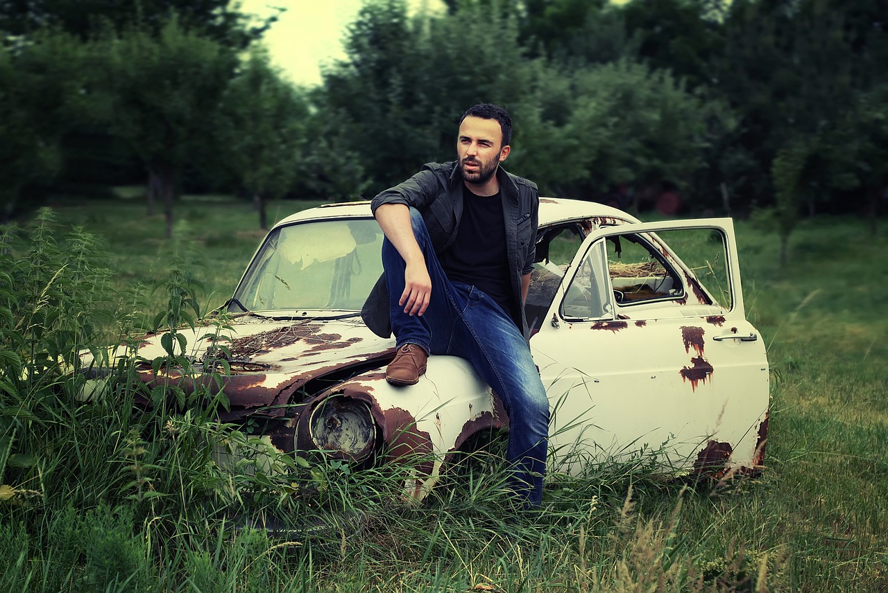 a man sitting on top of an old car in a field, an album cover, inspired by Nadim Karam, auto-destructive art, portrait mode photo, baris yesilbas, dressed in a white t shirt, background image