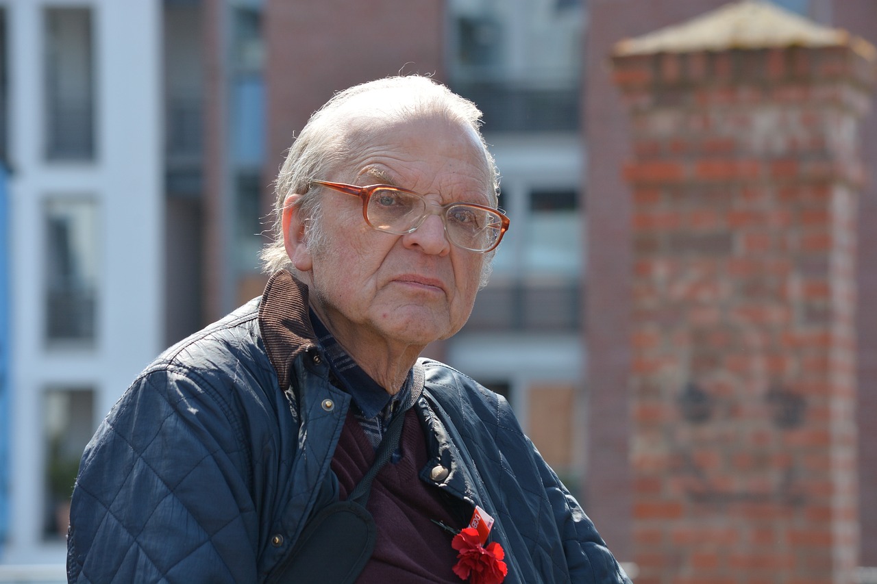a close up of a person wearing glasses and a jacket, a picture, by Jan Tengnagel, an 80 year old man, in an urban setting, red cloth around his shoulders, slightly sunny