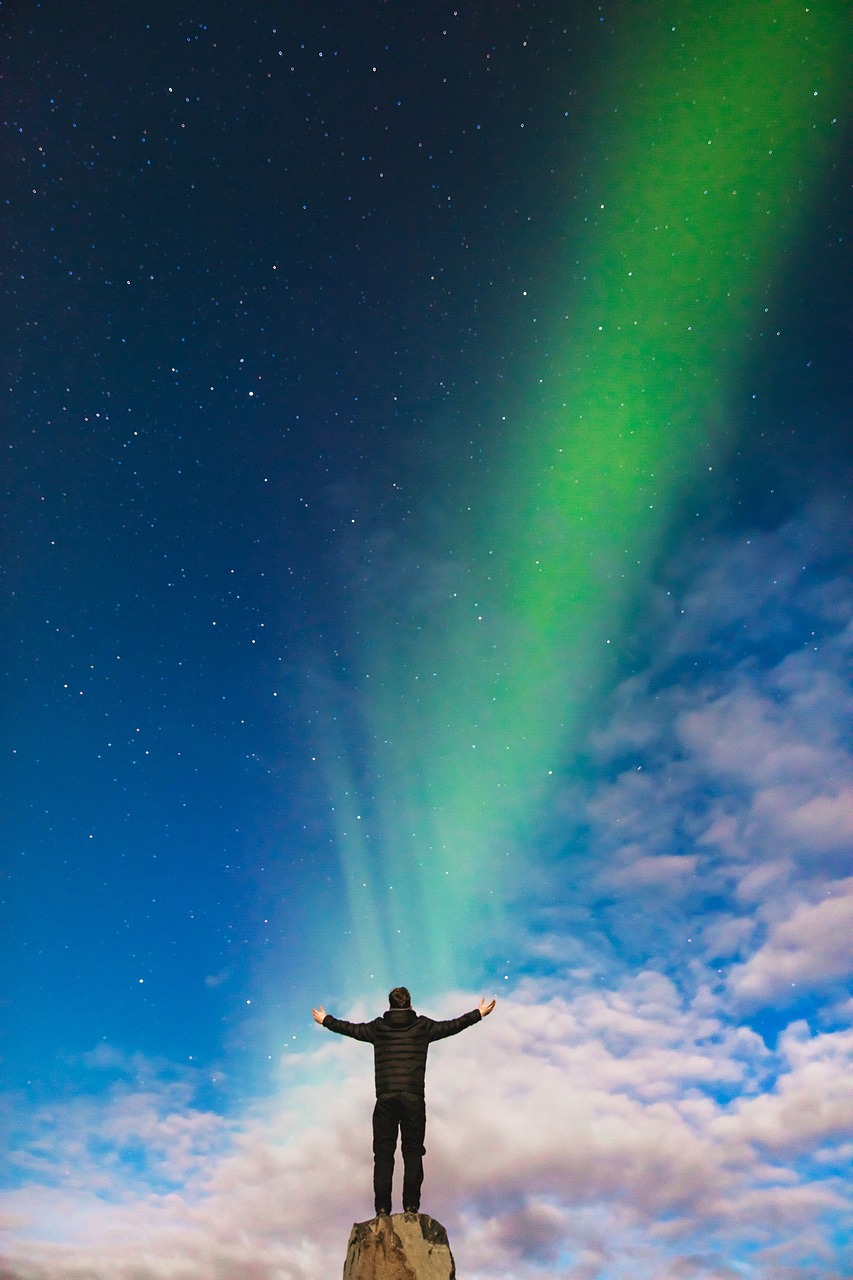 a man standing on top of a rock under a sky full of stars, by Ejnar Nielsen, blue and green rainbow fire, reykjavik, his arms spread. ready to fly, vertical wallpaper