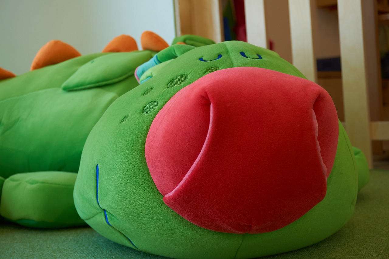 a green and red stuffed animal laying on the floor, inspired by Choi Buk, softplay, face center close-up, large comfy bed, soft draconic features