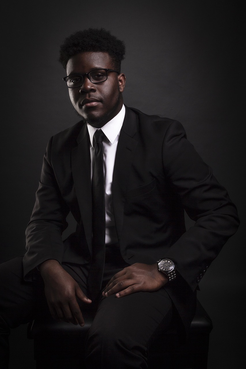 a man in a suit and tie sitting on a chair, an album cover, cg society contest winner, hurufiyya, dark black skin, shot at dark with studio lights, man with glasses, kevin hart