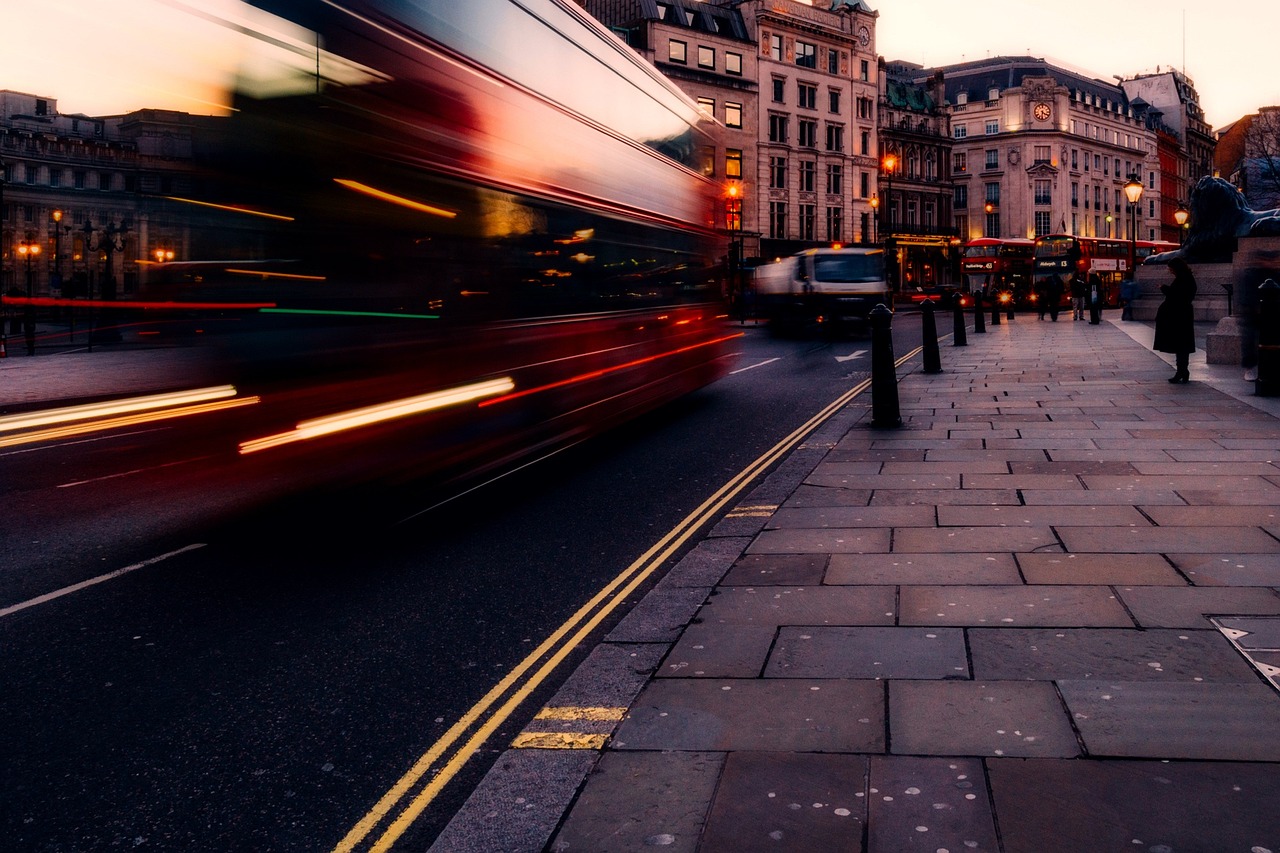 a double decker bus driving down a city street, a tilt shift photo, by Richard Carline, long exposure photo, evening at dusk, ((oversaturated)), street photography style
