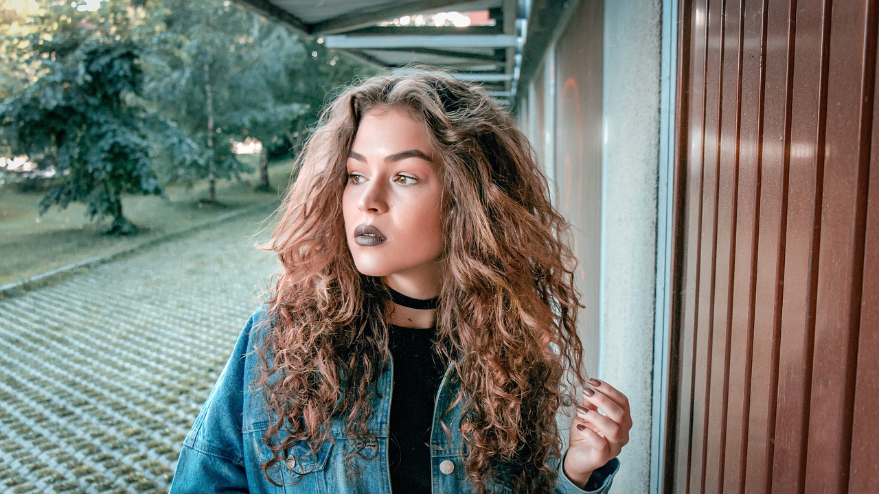 a close up of a person with long hair, inspired by Elsa Bleda, pexels contest winner, antipodeans, curly haired, wearing a jeans jackets, dark lipstick, standing outside a house
