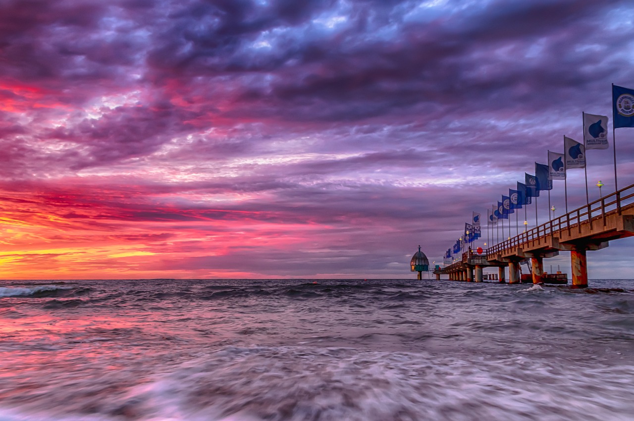 a pier filled with lots of flags next to the ocean, a photo, by Erik Pevernagie, fine art, colorful sunset!!, ultra wide, clouds and waves, redpink sunset