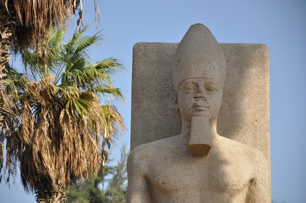 a statue of a man standing next to a palm tree, egyptian art, giant head statue ruins, ankh symbol around the neck, in modern era, outside