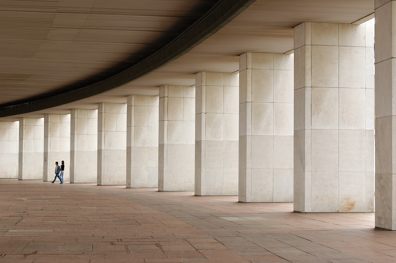 a couple of people that are walking through a building, a picture, inspired by David Chipperfield, unsplash, brutalism, colonnade, wikimedia commons, rock columns, 60mm