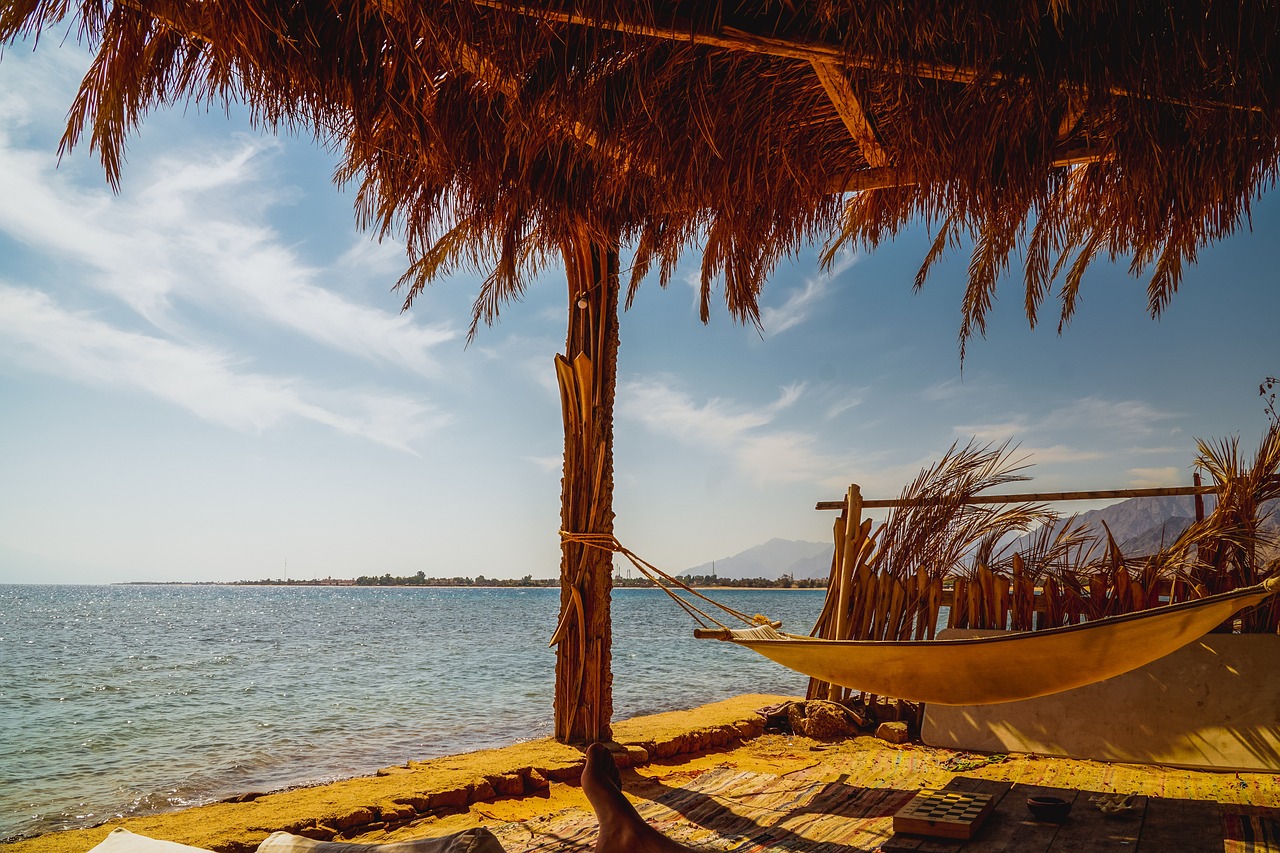 a hammock sitting on top of a beach next to a body of water, a stock photo, shutterstock, fine art, an amusement park in old egypt, shot on leica sl2, thatched roof, red sea