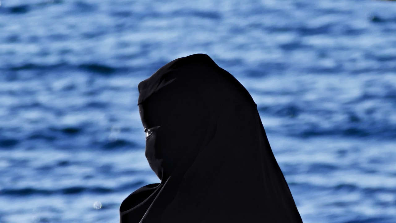 a person standing in front of a body of water, a picture, by Ibrahim Kodra, hurufiyya, veiled face, dark visor covering face, !female, mediterranean