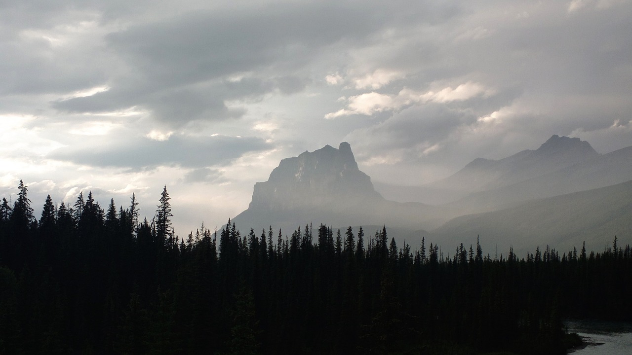 a view of a mountain range with a river in the foreground, tonalism, looks like a tree silhouette, banff national park, dark towering clouds, july 2 0 1 1