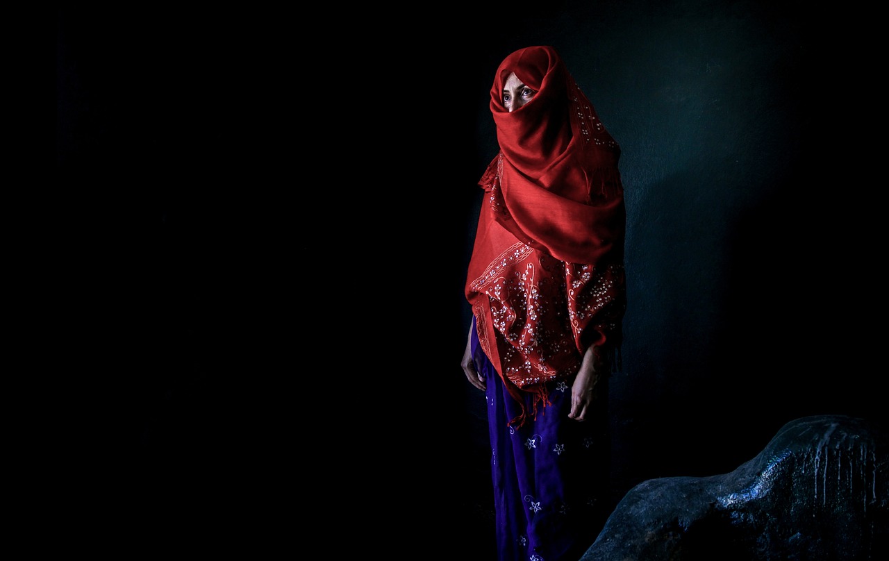 a woman in a red scarf standing next to a rock, a portrait, by Riza Abbasi, flickr, sony world photography awards, but the armor covers her face, standing in a dimly lit room, indigo