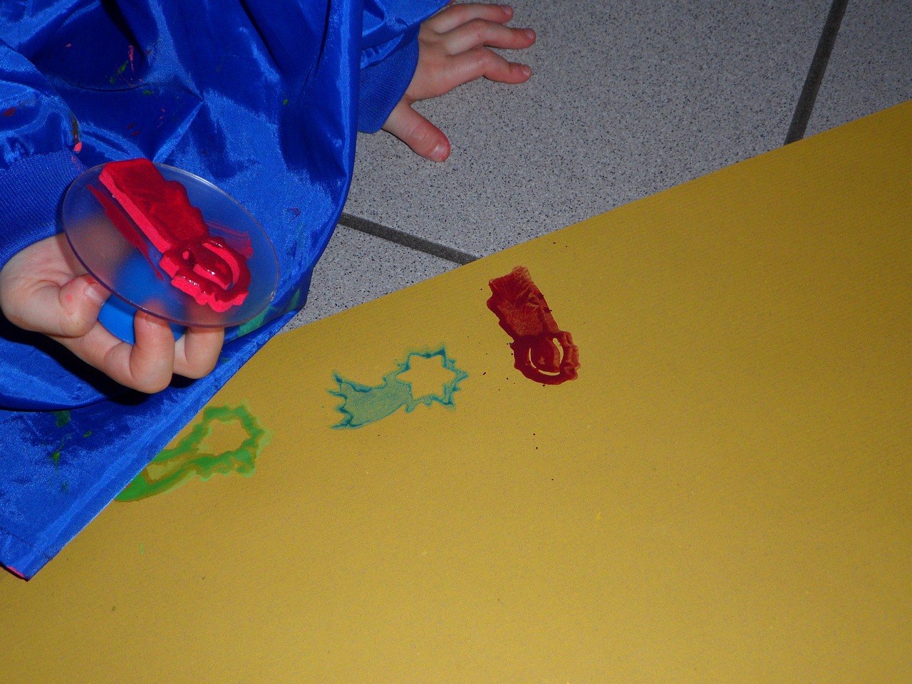 a close up of a child's hand holding a plastic object, a child's drawing, inspired by Lynda Benglis, action painting, wearing a red cape, acid pooling on the floor, shape of frogs, green blue red colors