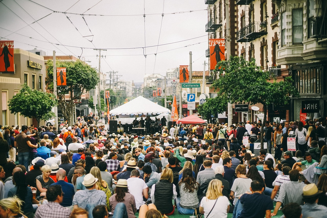 a crowd of people walking down a street next to tall buildings, by Matt Cavotta, happening, covered outdoor stage, sitting down, san francisco, big head on stage