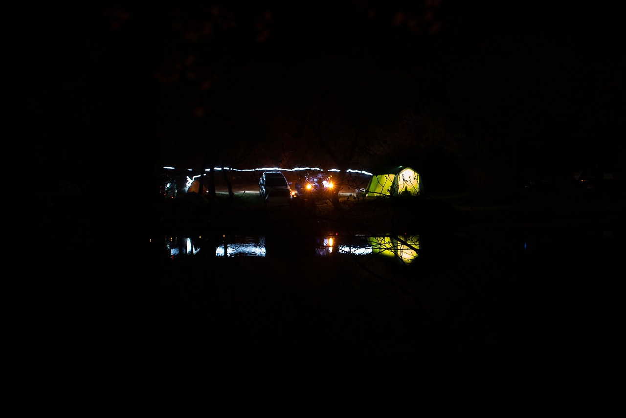 a boat sitting on top of a lake at night, a picture, by Richard Carline, tents, neon reflections in the puddles, brockholes, people angling at the edge