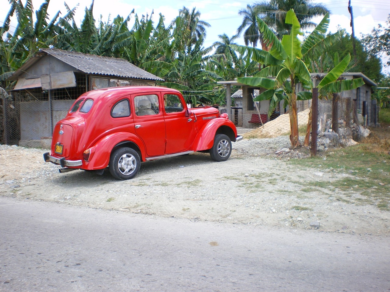 a red car is parked on the side of the road, by Ladrönn, flickr, cobra, jamaican, moskvich, hut, palm