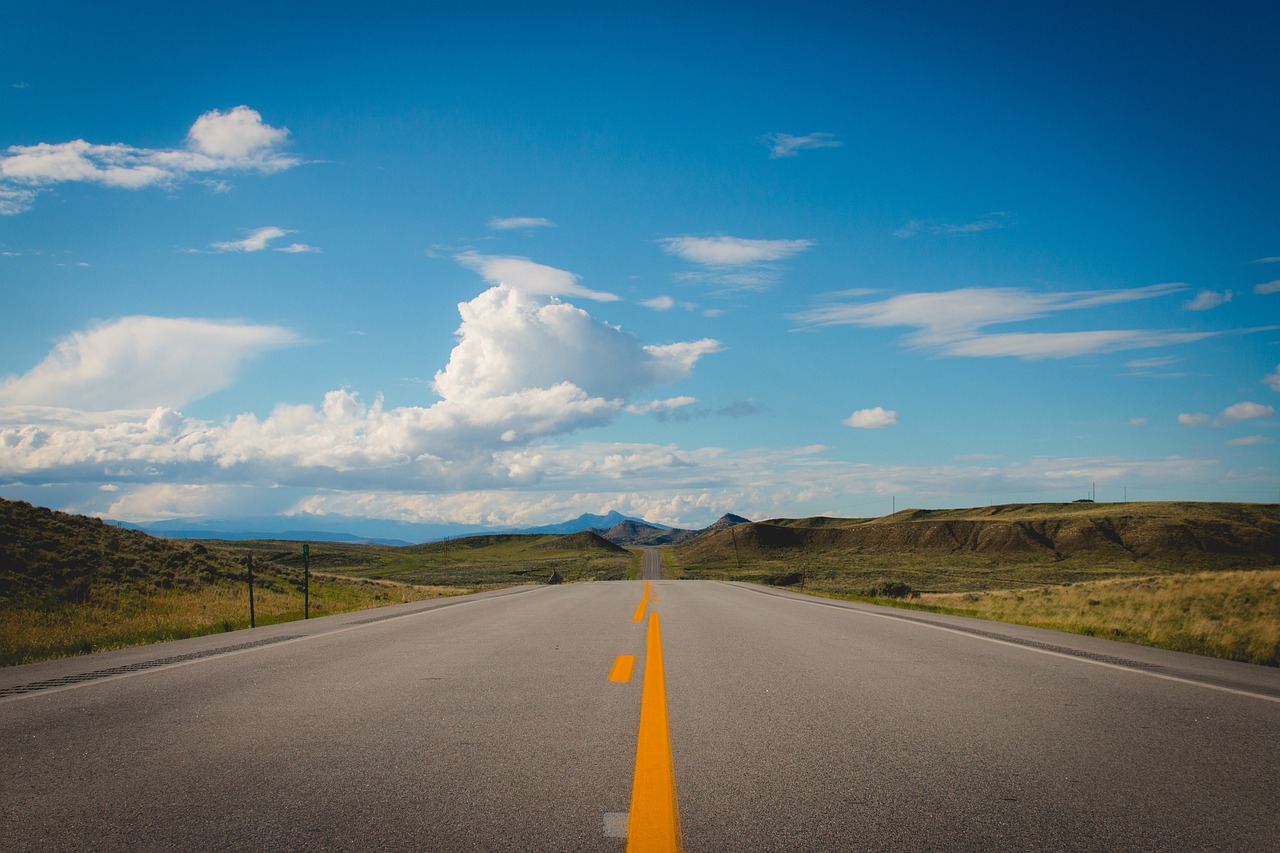 an empty road with mountains in the distance, a tilt shift photo, montana, big blue sky, long distance photo