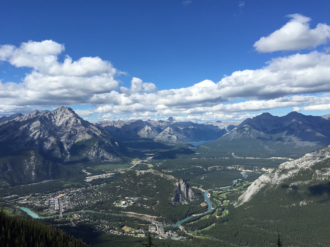 a view of a valley from the top of a mountain, by Brigette Barrager, happening, banff national park, entire city in view, blue river in the middle, looking partly to the left