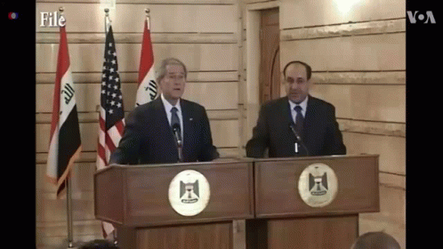 two men in suits and ties stand at a lectert with flags behind them