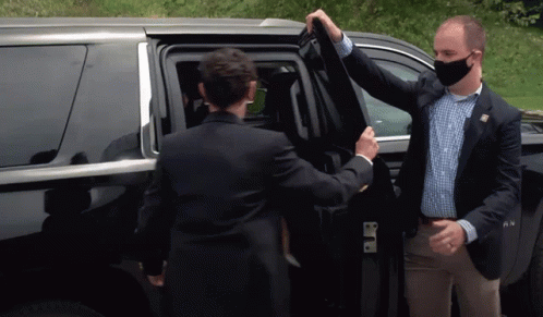a man in a suit and mask opening a car door while another man in a face mask holds up another mans tie