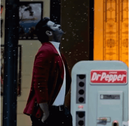 a man in a purple jacket looking up and to the sky next to a machine with a dr pepper sign