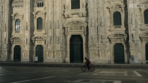 a guy on a bicycle is passing by a very large building