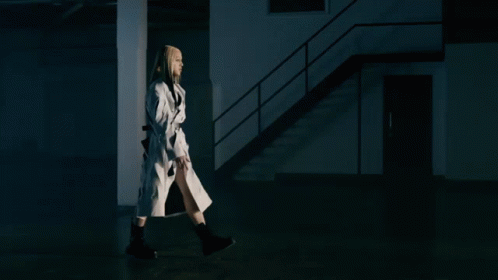 woman in a white trench coat walking by a staircase at night