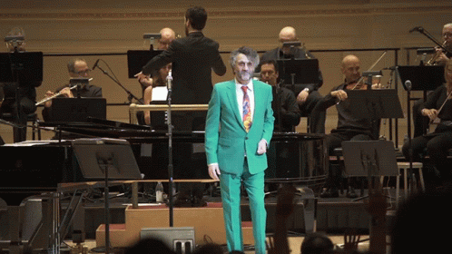a man in a suit with clown make - up and orchestra behind