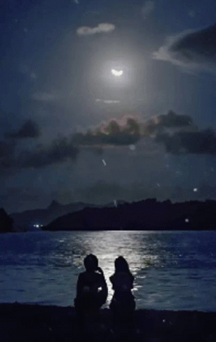 two people standing on a beach with the moon in the distance