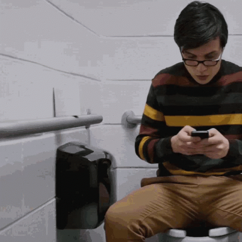 a young man sitting on a toilet looking at his phone