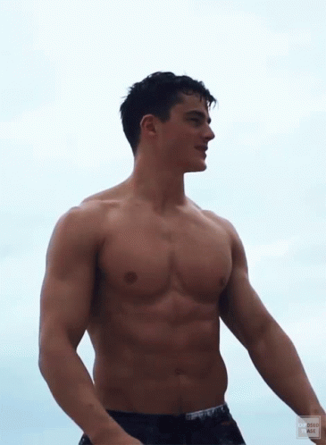 a shirtless male stands on the beach at sunset