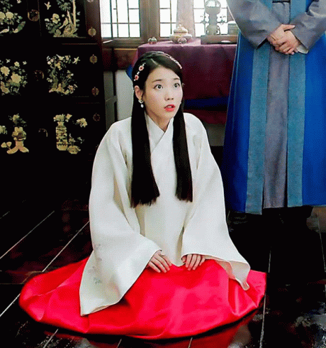 an oriental woman with long black hair, white and blue dress and a crown sits on the floor