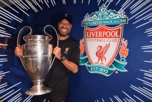 man holding silver trophy in front of wall with liverpool logo