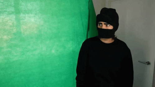 a person wearing a black mask stands by a green wall