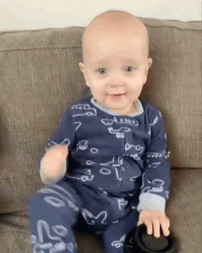 a baby in pajamas on a couch with one leg out and a black object in the other hand
