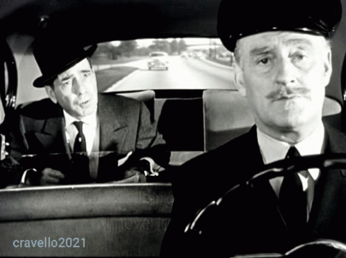 the driver in the cab of a car wearing a fedora and the other gentleman with a mustache