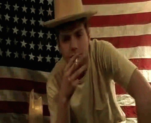 young man with fedora and cigarette in front of flag