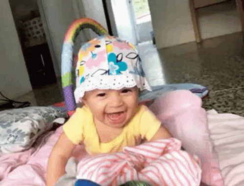 an infant with a hat and blanket sits on a bed and smiles