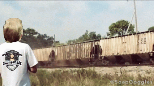 a boy in an unisex t - shirt watches as a train goes by